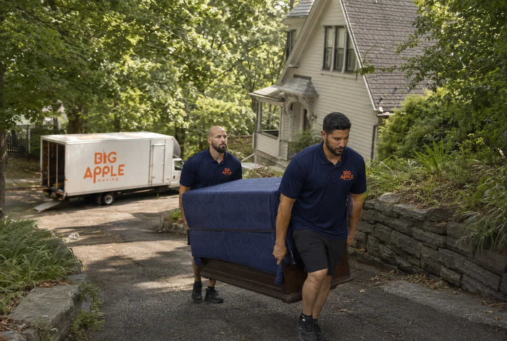 Two Big Apple Moving movers carrying a blanket-wrapped dresser down a sloping driveway to a waiting truck