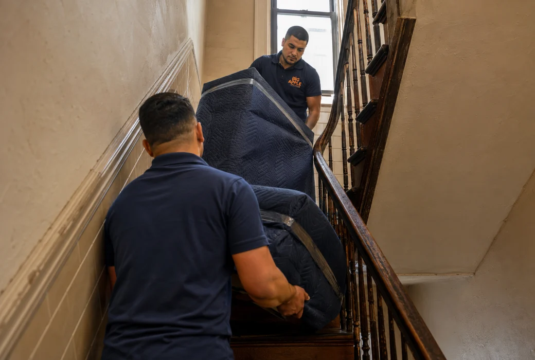 Two Big Apple Moving movers carrying a wrapped armchair up a narrow New York City walk-up staircase